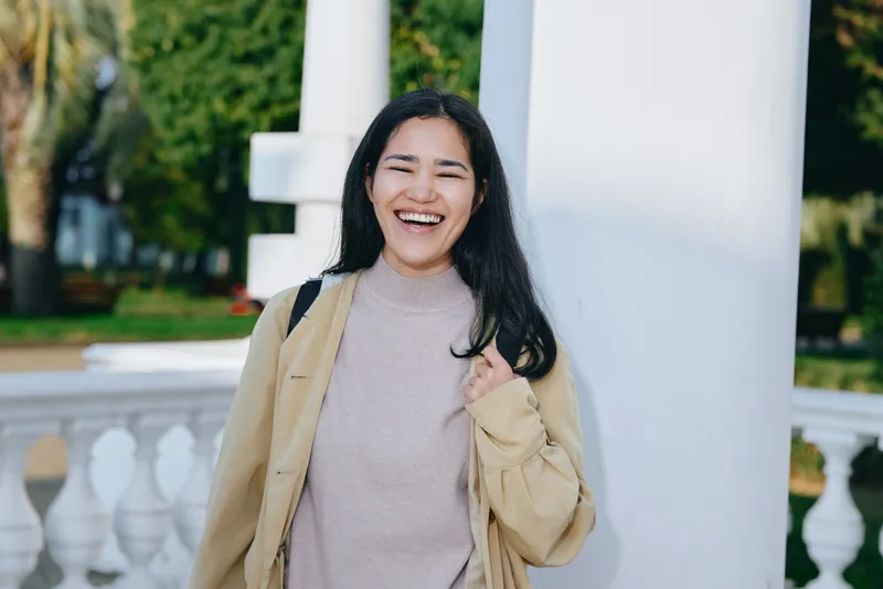 Woman in a casual jacket standing outdoors by white columns, smiling warmly in daylight, conveying a friendly, relaxed street style and positive mood for lifestyle use.