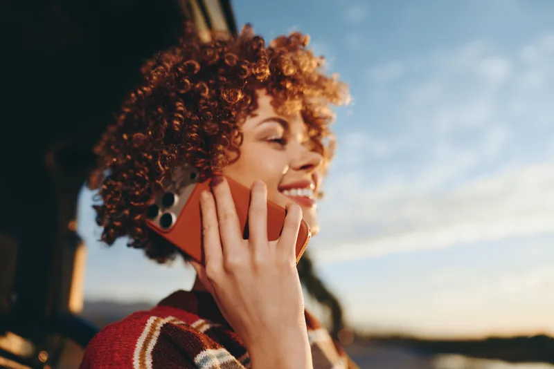 Woman with curly red hair smiling while speaking on a smartphone outdoors at sunset, warm light and soft bokeh, lifestyle communication, confident mood, evening calm routine today