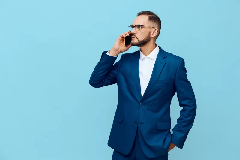 A business professional man stands against a solid blue background, projecting confidence and focus as he speaks on a mobile phone during a strategic work moment.