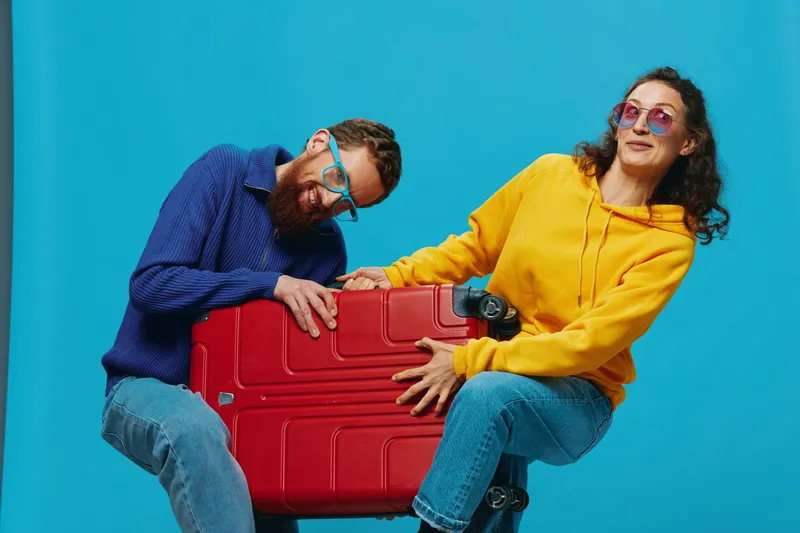 Женщина и мужчина улыбаются, держат чемоданы, причем желтый и красный чемоданы весело улыбаются, на синем фоне