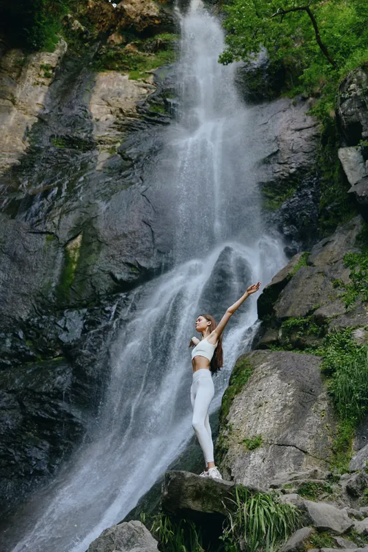 woman near waterfall standing on rock outdoors surrounded by lush greenery and rugged cliffs in bright natural light