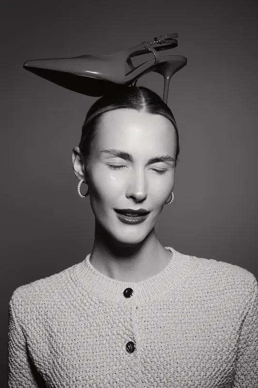White woman with fair skin and closed eyes balances high heel shoe on head in studio. Concept of fashion, elegance, and creative portrait in black and white.