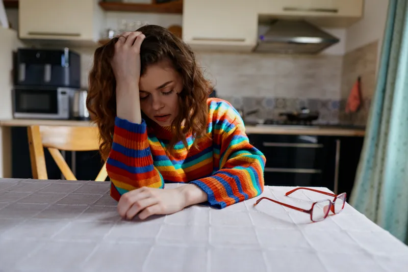 Young woman sits at a kitchen table indoors, resting her head on her hand in a cozy home moment, wearing a colorful striped sweater and round glasses.