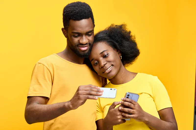 Young couple enjoying a moment together while browsing on a smartphone against a vibrant yellow background, showcasing love and connection in casual outfits