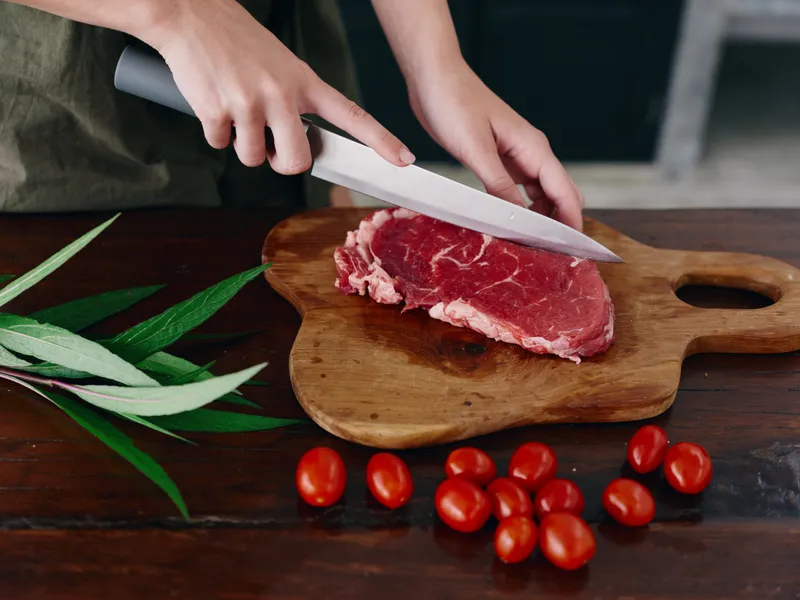 Женщина с ножом в руке нарезает свежий стейк из мяса для запекания на кухне
