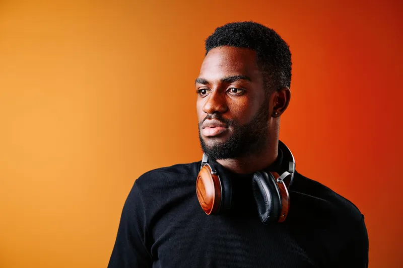 Young Black man wearing headphones against a vibrant orange background, expressing contemplation and modern style, ideal for music or lifestyle concepts