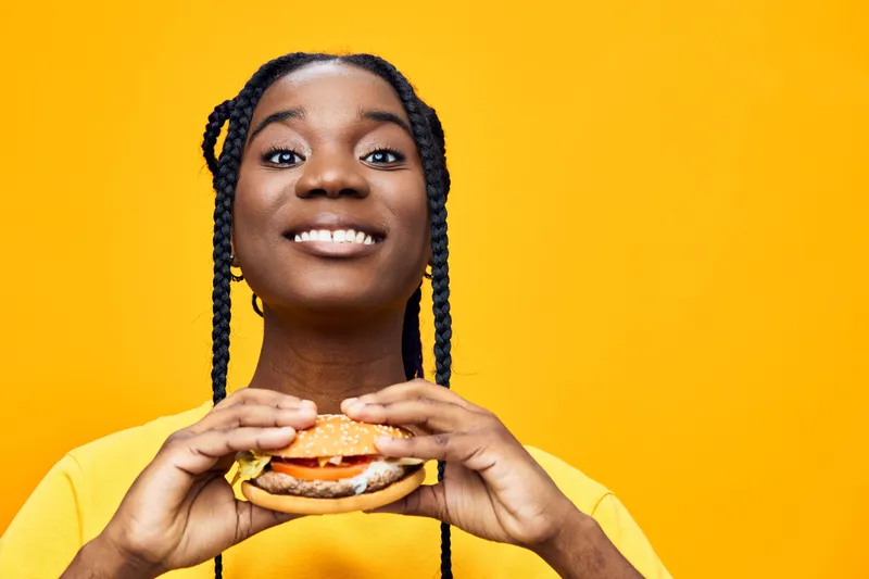Happy young woman with braids holding a delicious burger, wearing a bright yellow shirt, on a vibrant yellow background, showcasing healthy eating and joy