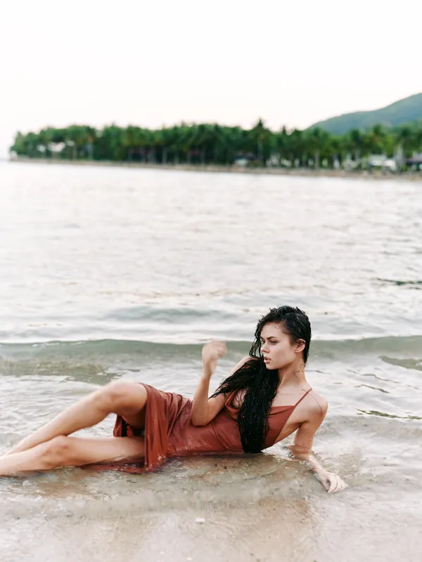 A woman lies semi submerged on a shallow shoreline wearing a flowing brown dress, posing gracefully as gentle waves lap at her side.