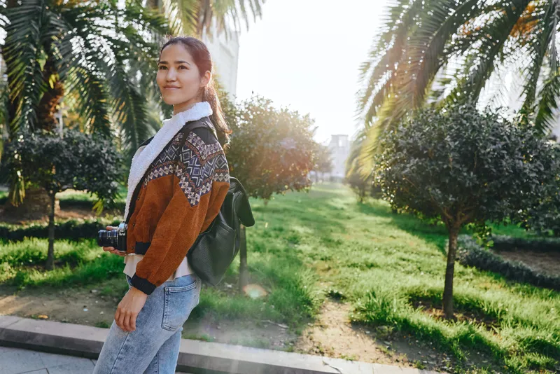 Woman with backpack walks along a sunlit park path, casual fashion and scarf fluttering, greenery and trees in the background, candid outdoor moment captured during golden hour.