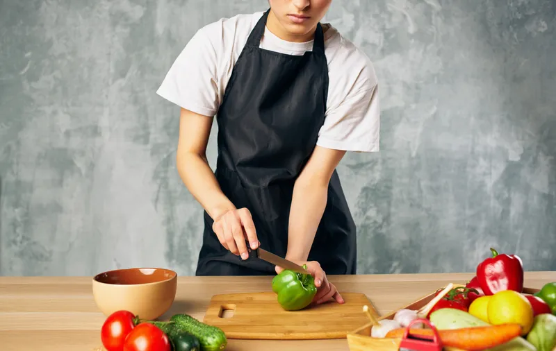 Женщина в черном фартуке нарезает овощи на кухне, готовит свежую еду