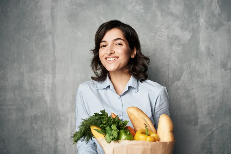Весёлая брюнетка с пакетом продуктов, в синей рубашке, на фоне овощей и тёмного фона