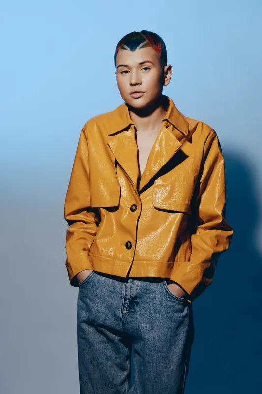 Young woman with short colorful hair wearing a stylish yellow jacket and casual jeans posing against a blue gradient background, conveying modern fashion and confidence.