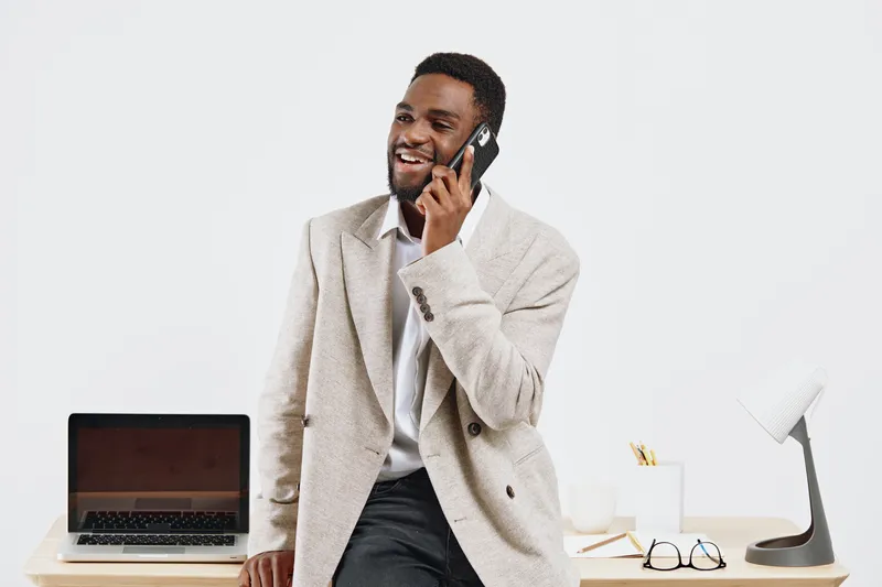 Smiling man wearing beige blazer and white shirt talking on mobile phone, leaning on desk with laptop and stationery, in bright modern office interior. Business people lifestyle concept