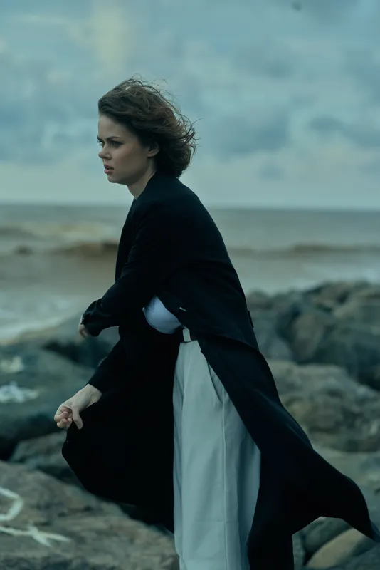 Wind swept coast scene with a woman in a flowing coat on a rocky shoreline, moody sea in background, emphasizing motion, drama, and outdoor resilience