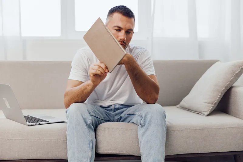 Thoughtful man sitting on a couch, reading a notebook at home, featuring modern decor and a warm atmosphere. Ideal for showcasing creativity and introspection.
