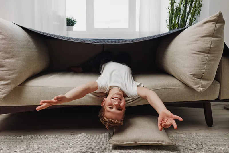 Happy playful boy lays upside down under beige couch in bright living room, smiling and reaching out with arms. Concept of childhood fun, home play, and carefree moments.