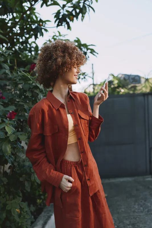 Casual woman outdoors in a rust colored outfit, curly hair, relaxed pose, sunlit moment, soft breeze, natural colors, greenery background, lifestyle fashion scene, confident expression