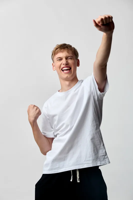 Joyful celebration and victory vibe as a young man raises a fist in triumph, energetic posture in a white t shirt, bold stance, bright studio background conveying motivation and success