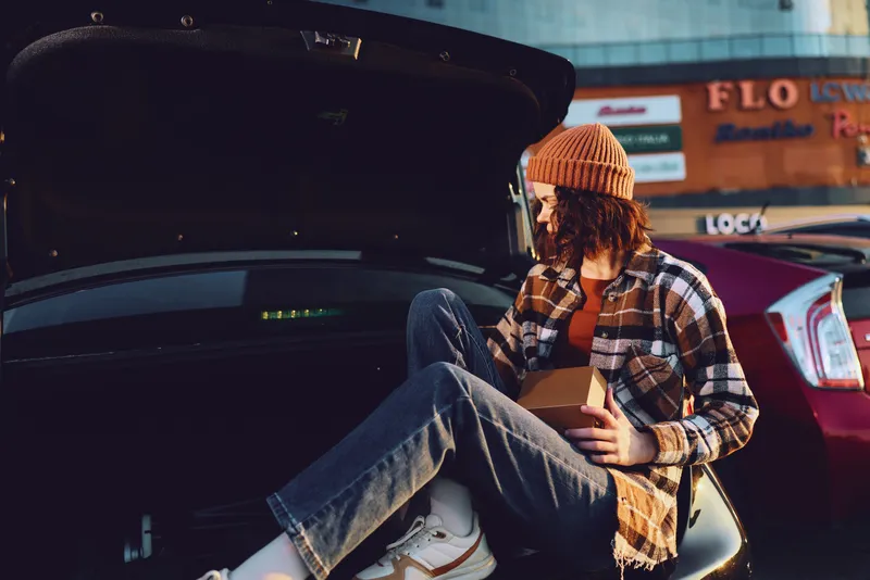Man car trunk takeaway beanie flannel urban night portrait of a candid lifestyle moment: young man eating takeout on an open car trunk with authenticity, golden hour glow, emotional storytelling.