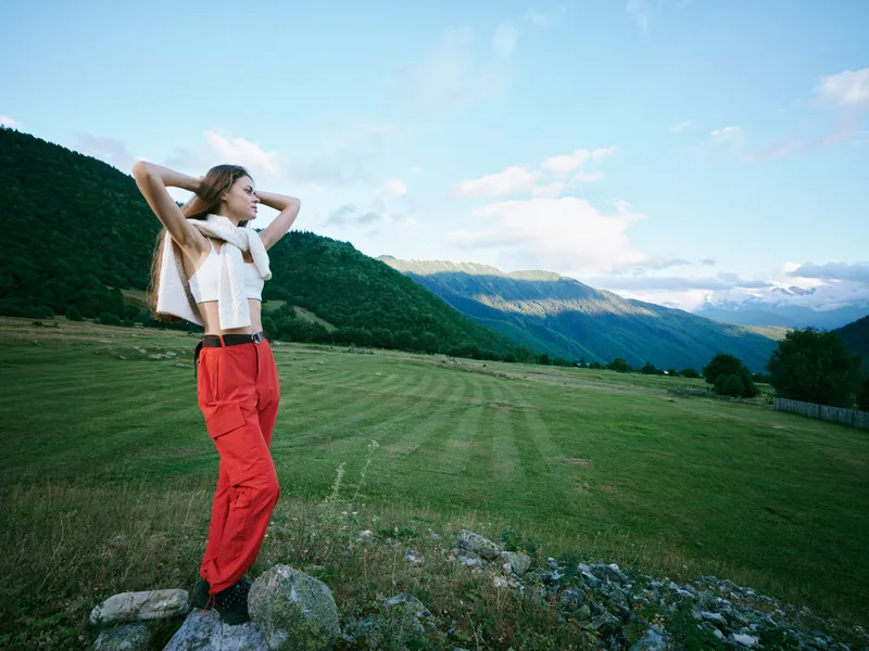 Woman in red trousers and scarf stands on rocks in an open field with mountains, capturing a moment of outdoor freedom and scenic landscape