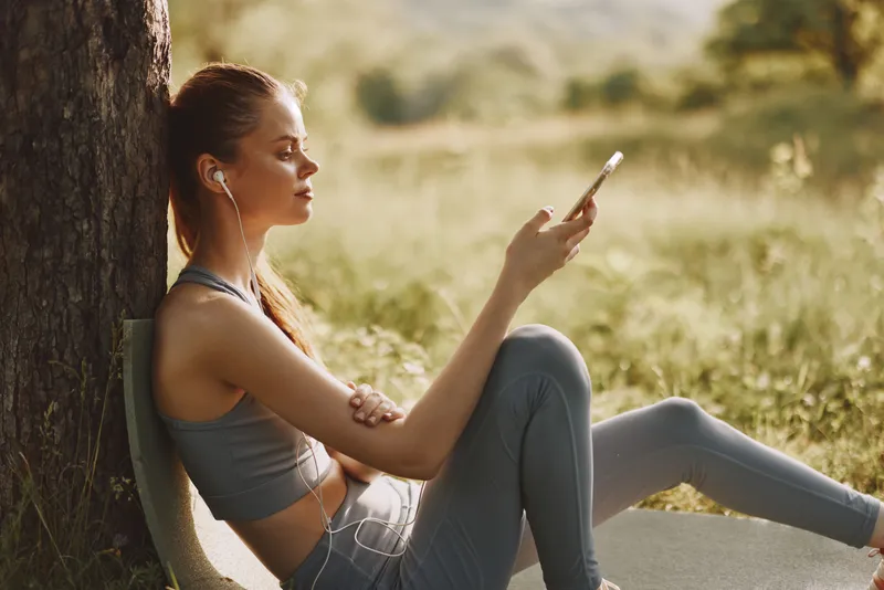 Young woman enjoying music outdoors in activewear under a tree, feeling relaxed and connected with nature during summertime, providing inspiration for wellness