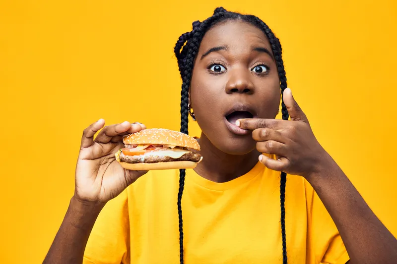 Happy young African American woman enjoying a delicious burger against a vibrant yellow background, expressing joy and surprise with playful gestures