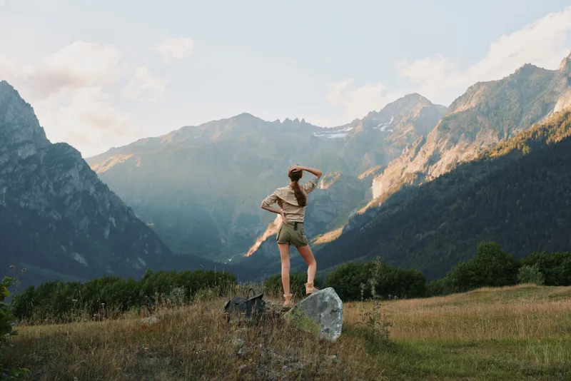 Young woman stands on a rock in a sunlit valley, arms raised in triumph as distant mountain peaks glow under a clear blue sky, wearing beige tank top and light shorts, embodying freedom