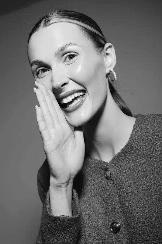 Smiling young Caucasian woman with light skin and glossy hair holds hand near mouth as if whispering, wearing textured jacket in studio. Concept of communication and happiness.