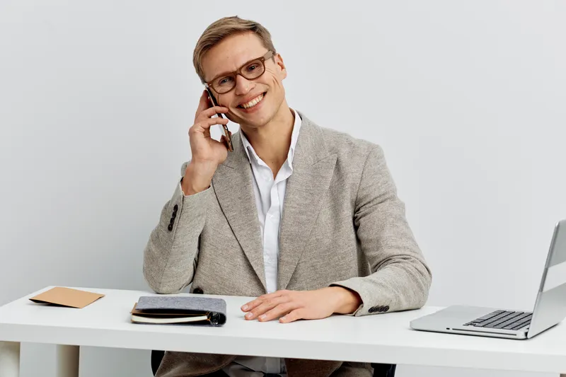 Smiling man wearing glasses, beige blazer, white shirt, talking on mobile phone, sitting at office desk with laptop, notepad, and pen, cheerful indoor workspace, business communication concept.