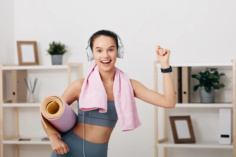 Woman in athletic wear with headphones, holding a yoga mat and towel, celebrating a workout at home with an energetic pose and bright shelves in the background.