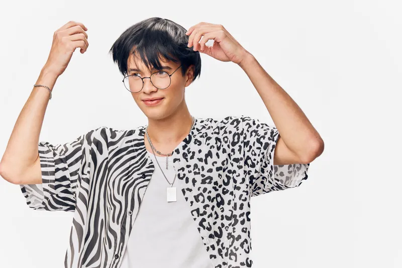 young man with stylish glasses and zebra-leopard print shirt adjusting hair, trendy fashion, casual, confident expression in white studio setting