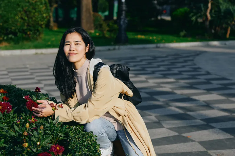 Woman kneeling beside flowers in a park, outdoor scene with backpack, casual attire, gentle smile, sunlight among greenery, calm nature moment suitable for stock photography, woman enjoying serene