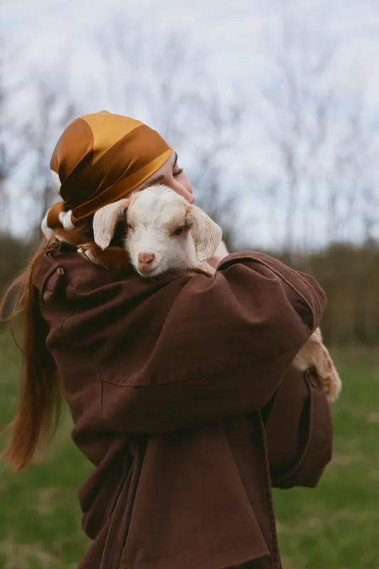 Young woman wearing a brown jacket and headscarf gently hugs a small white baby goat outdoors on a cloudy day in a peaceful countryside setting.