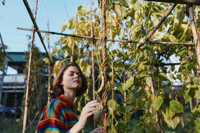 Woman in a striped sweater and straw hat, young adult portrait in an urban garden among climbing plants, standing and touching vines with contemplative expression in soft sunlight lifestyle scene.