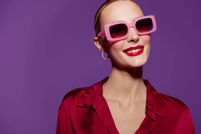 Smiling woman with red lipstick wearing pink sunglasses and red blouse poses against purple background. Concept of fashion, style, beauty, and confident female portrait.