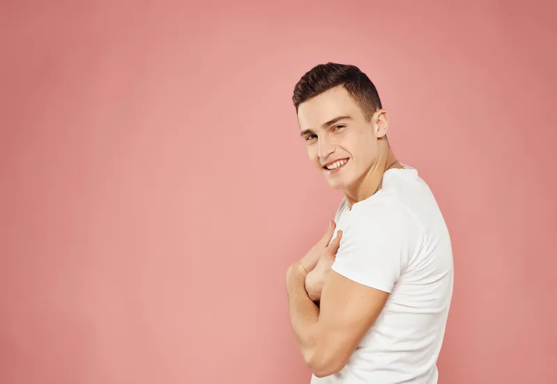 Весёлый мужчина жестикулирует руками, одетый в белую футболку на розовом фоне