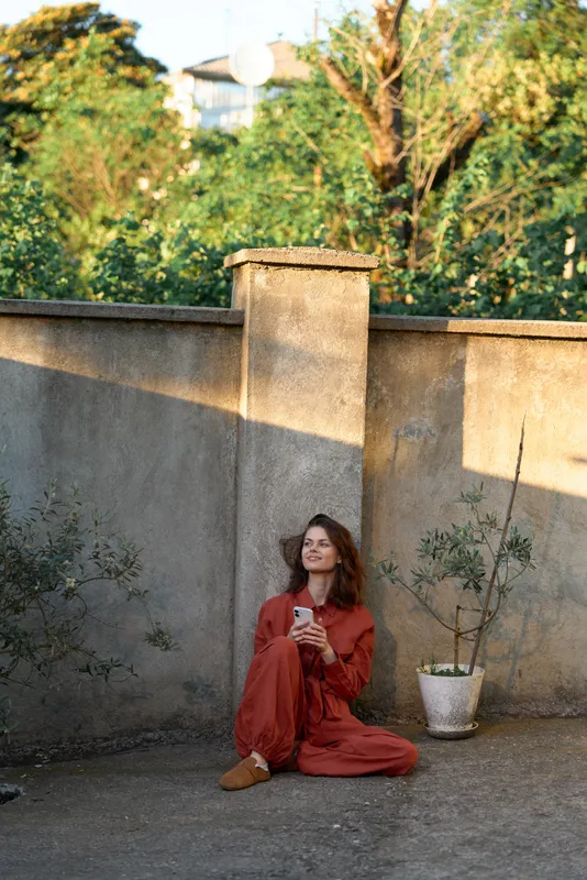 A woman seated on the ground in a rust red jumpsuit, outdoor setting beside a concrete wall, holding a smartphone. A calm, casual lifestyle moment in a daytime sunlit garden