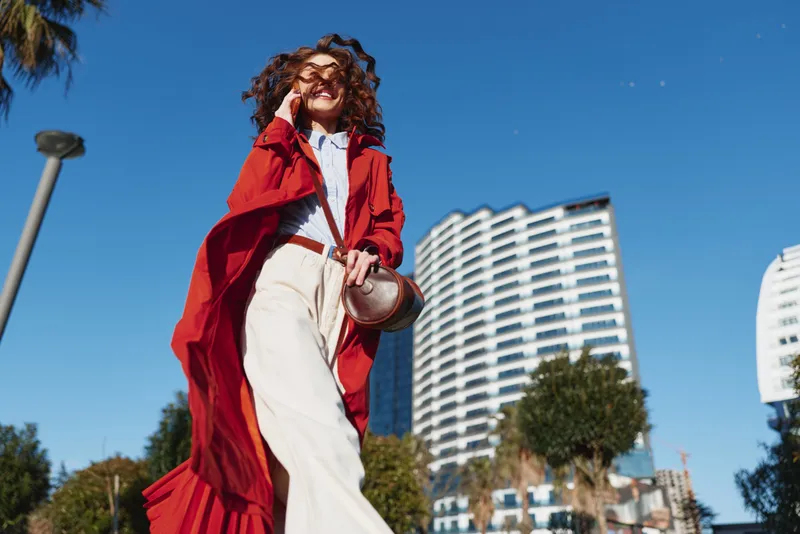 Smiling woman in a flowing red cape walks through a sunny urban street, showcasing bold fashion, wind swept movement, and contemporary style in daylight.