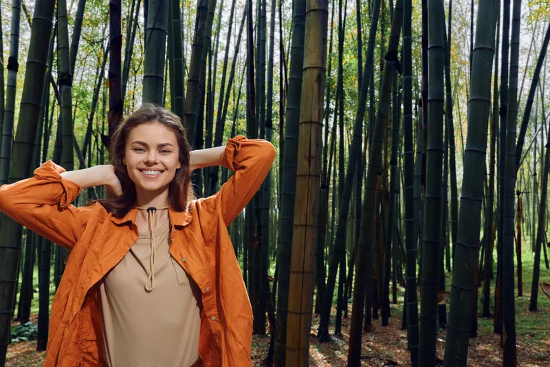 Woman smile bamboo forest portrait nature outdoor happy model trees, smiling woman with hands behind head in orange jacket, relaxed and serene amid tall green bamboo and soft sunlight.