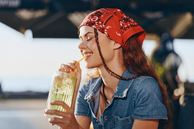 lifestyle woman in modern boho-western denim street style enjoying a refreshing drink, smiling with eyes closed, film color tones creating warm vibes outdoors