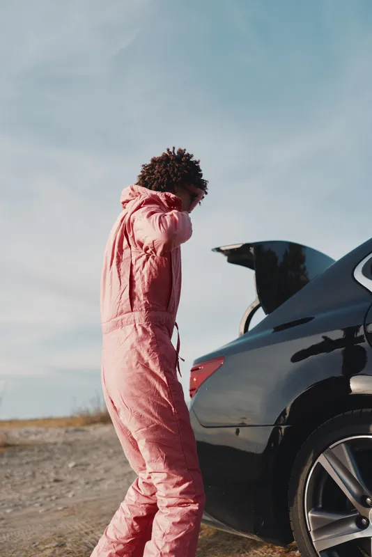 Car trunk open, outdoors scene with a person wearing a pink jumpsuit by a road in a desert, preparing belongings under a clear sky and bright sunlight.