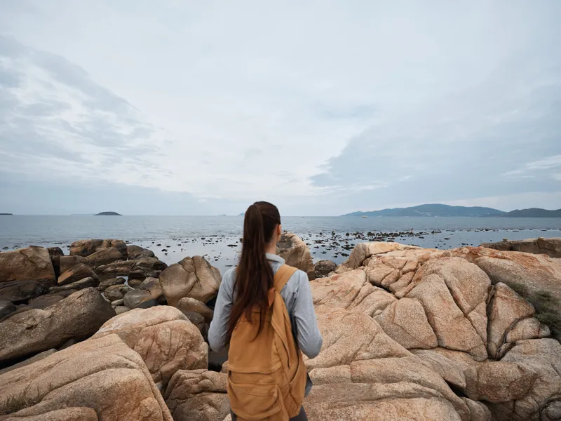 Young woman exploring rocky coast during overcast day, wearing casual clothes and backpack, embodies adventure and connection with nature