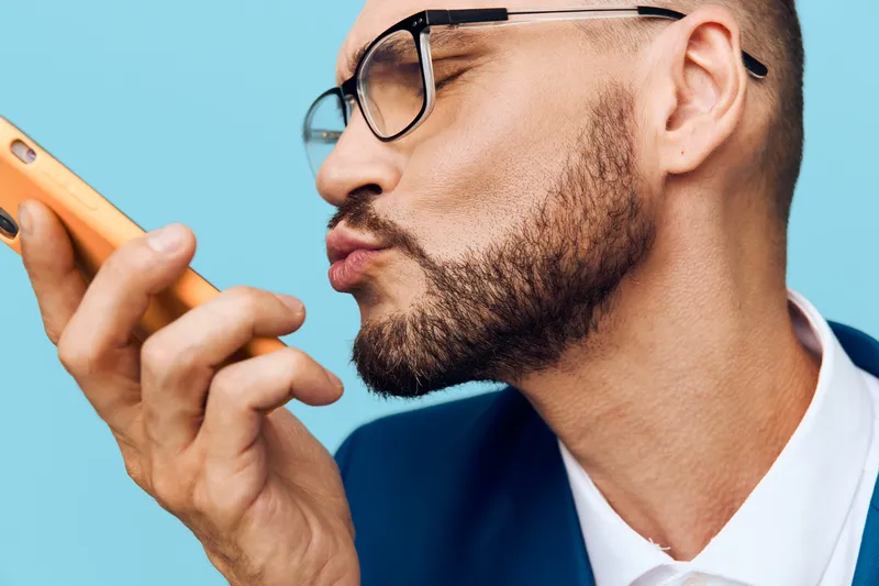 A professional man in business attire stands against a solid colored backdrop, projecting confidence and focus. He peers at a device with careful emotion, highlighting modern work life and strategic