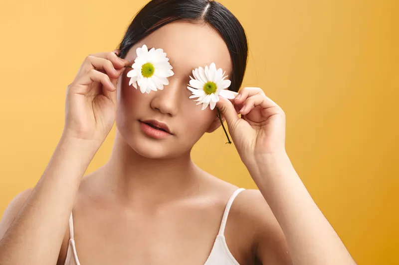 Asian woman is seen in a bright studio holding white daisies over her eyes, creating a playful, whimsical portrait with soft lighting and a calm, creative mood.