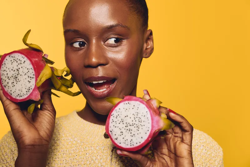 Close-up of a young black woman with dark skin holding two halves of a dragon fruit against yellow background. Concept of healthy lifestyle, exotic fruit, and natural beauty.