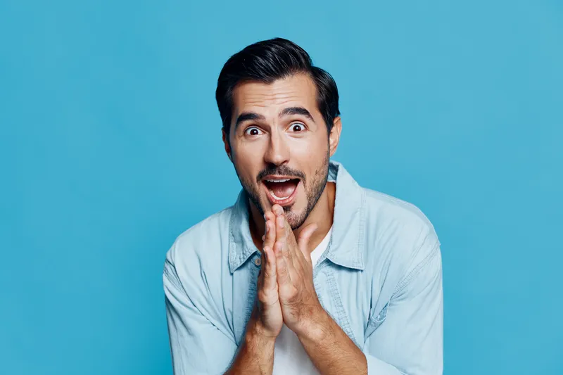 Happy surprised man with dark hair wears light shirt, gestures joyfully with hands near face, isolated on vibrant blue background. Excitement and enthusiasm concept.