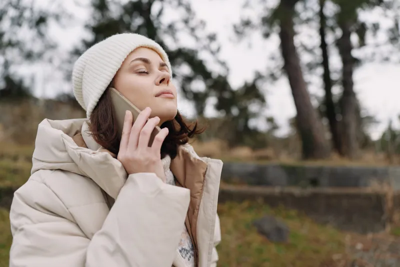 Woman outdoors wearing a coat and beanie talking on a phone in a tranquil park, capturing a candid lifestyle moment with autumn light and calm mood, ideal for fashion, travel or everyday use