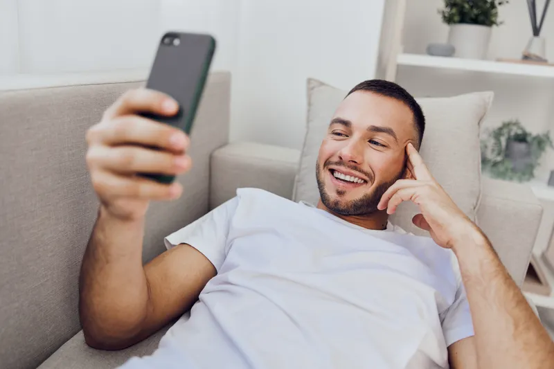 Happy man relaxing at home while using a smartphone, showcasing modern digital communication and leisure time in a cozy environment.