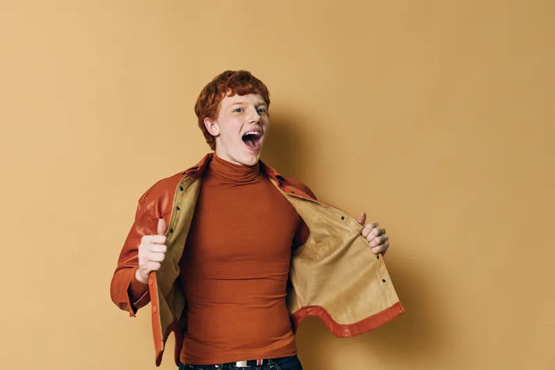 Young man with curly hair wearing brown turtleneck and leather jacket posing excitedly with open coat on beige background. Happy expression and casual style.