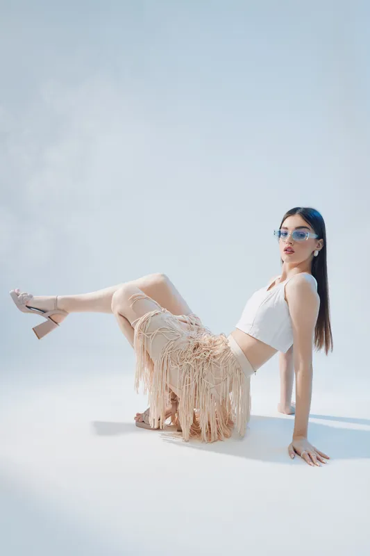 Young woman in fashionable fringe skirt and white crop top poses seated on floor against light background. Concept of woman beauty fashion with stylish accessories and modern look.